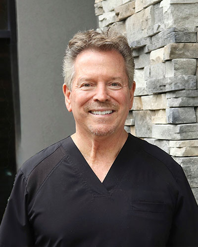 The image shows a man standing next to a stone wall with a smile on his face.