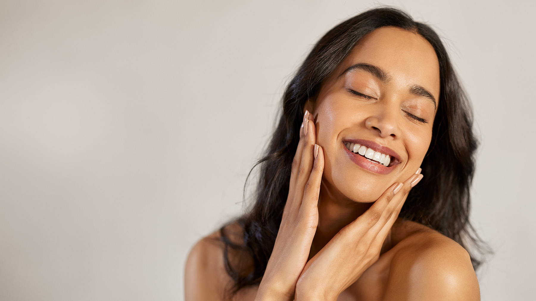 A woman with closed eyes, smiling slightly, looking upwards while applying skincare product to her face.