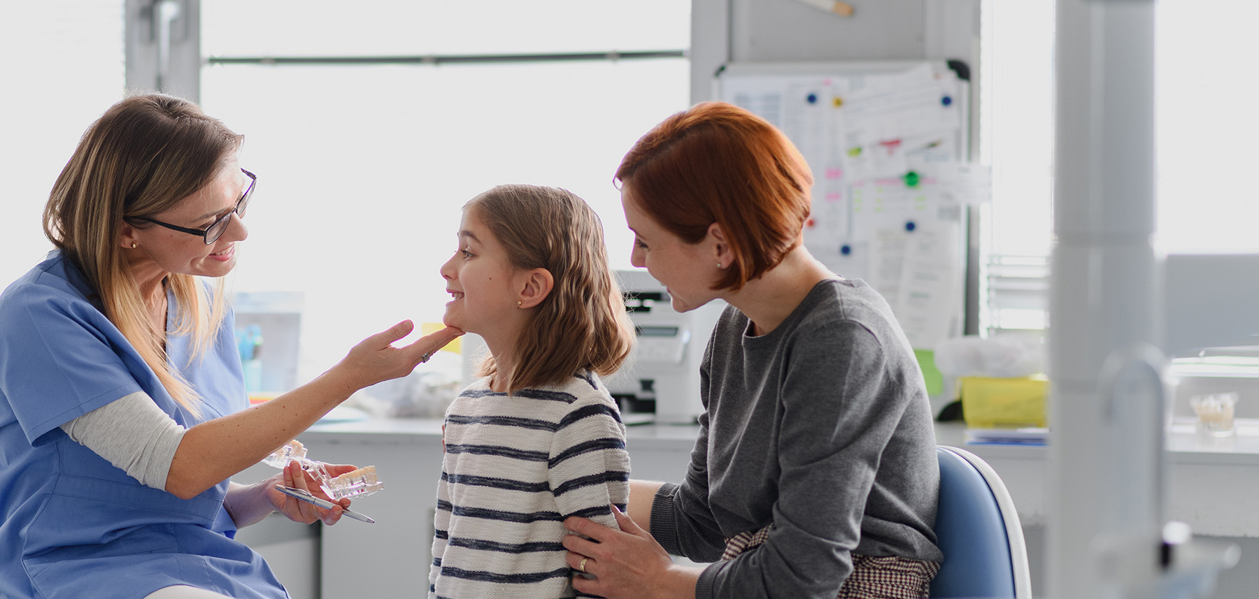 The image shows a group of people in a medical setting, with a woman holding a child s face steady while another person appears to be performing a dental procedure on the child.
