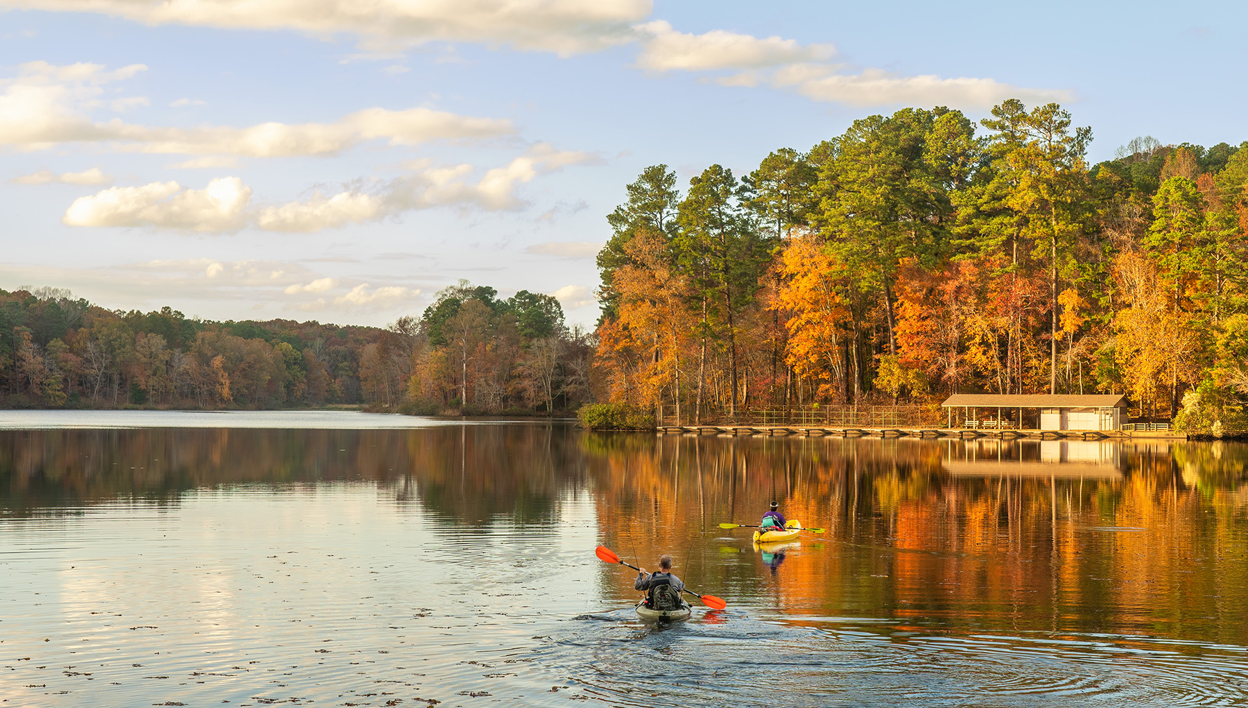 The image depicts a tranquil scene with a person kayaking on a calm lake surrounded by trees and autumn foliage, with a clear sky above.