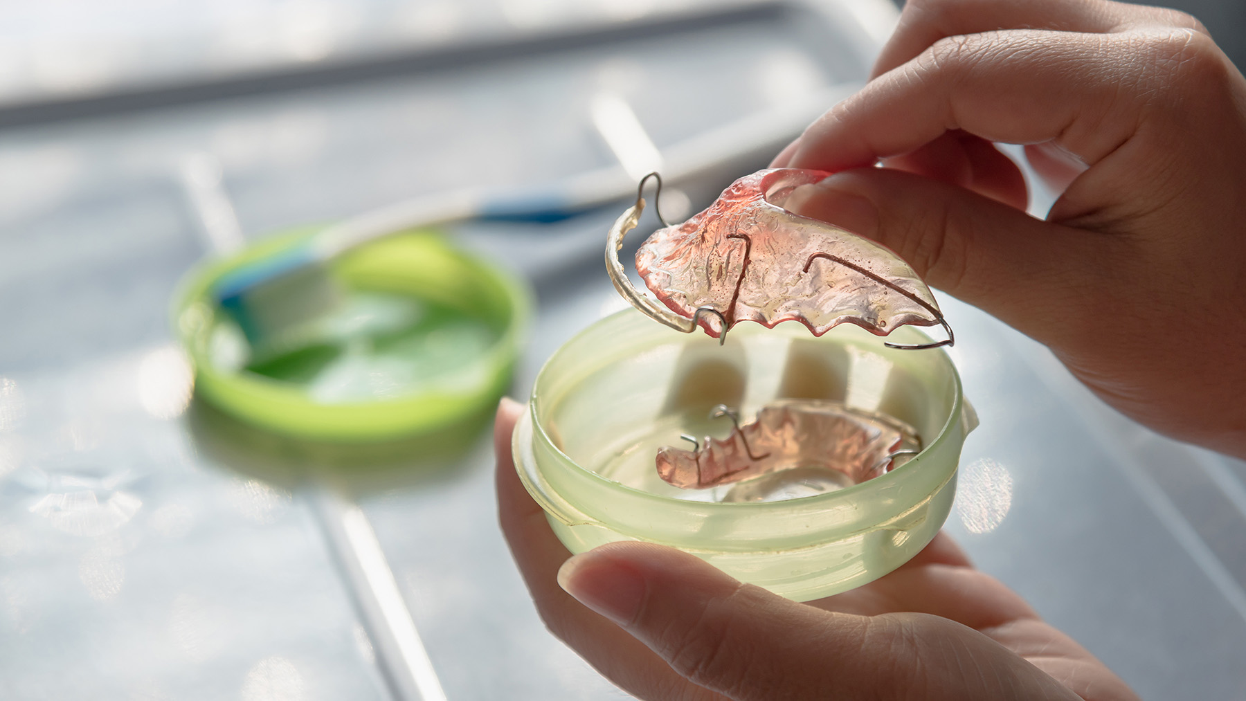 The image features a hand holding a small green plastic container with an intricate pink butterfly design inside, placed on a table with various items around it.