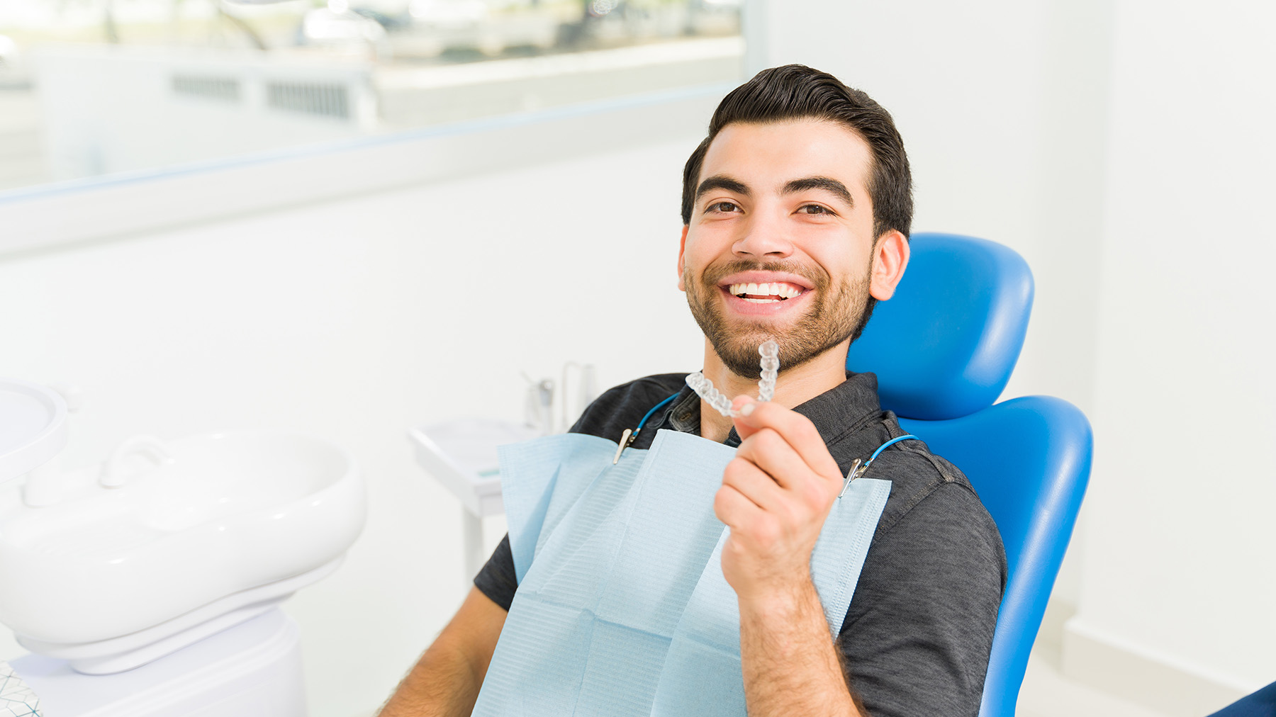 A smiling man holding a toothbrush in front of his mouth while sitting in a dental chair with dental instruments around him, likely in a dental office setting.