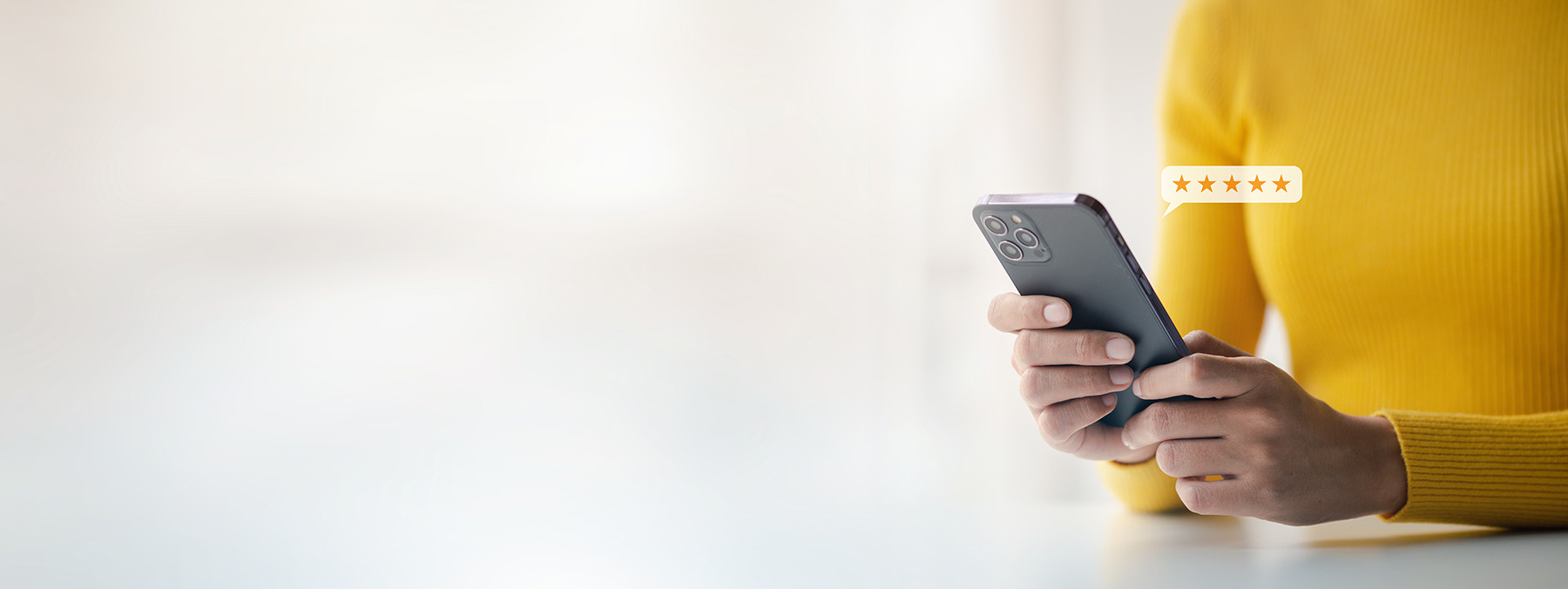 The image shows a person using a smartphone with a focus on their hands holding the device, which is placed on a surface. The background is blurred but appears to be an indoor setting with warm lighting.