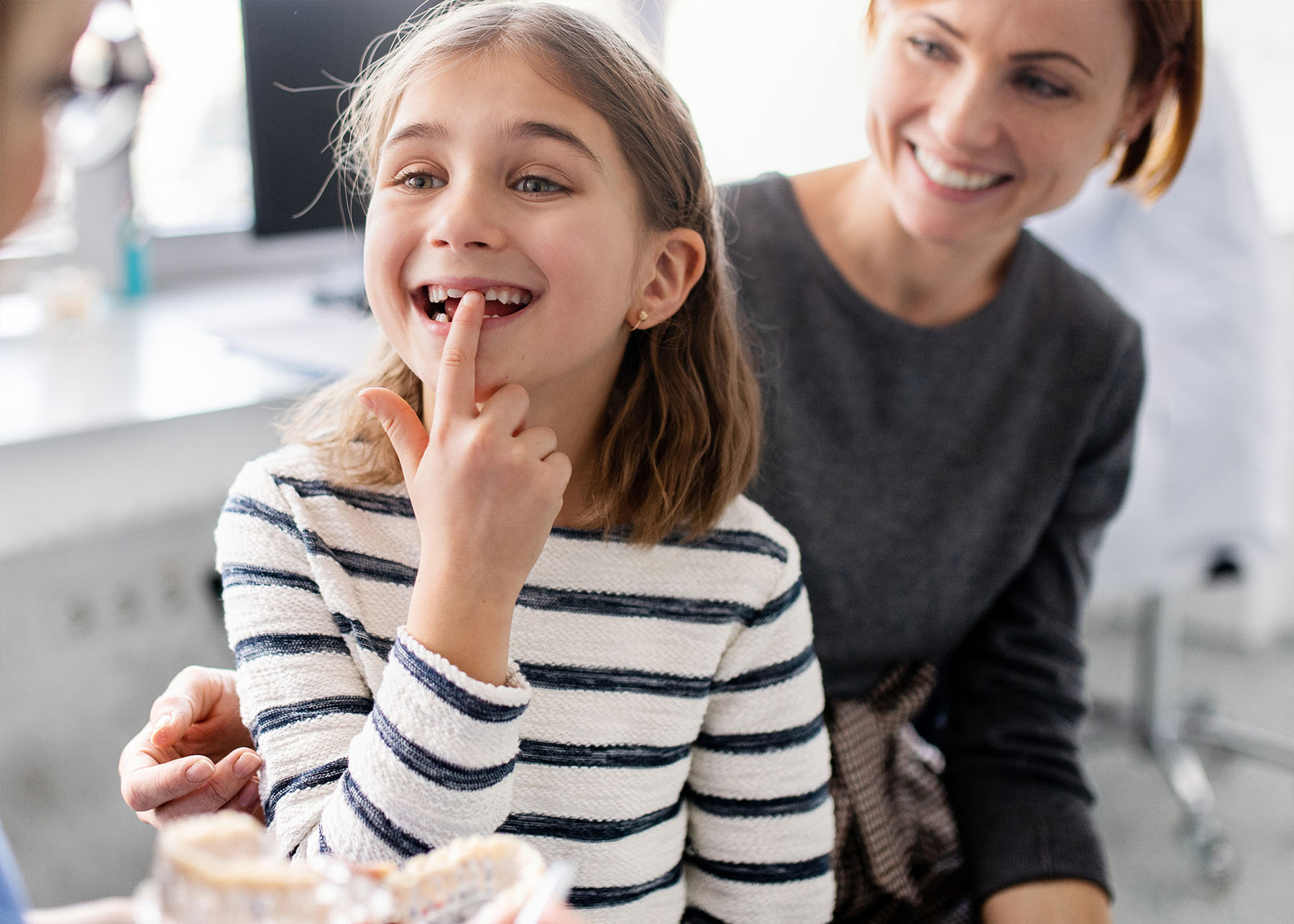A young girl with her mouth open is sitting in front of an adult woman, both are indoors, likely in a dental office setting, with various dental equipment visible around them.