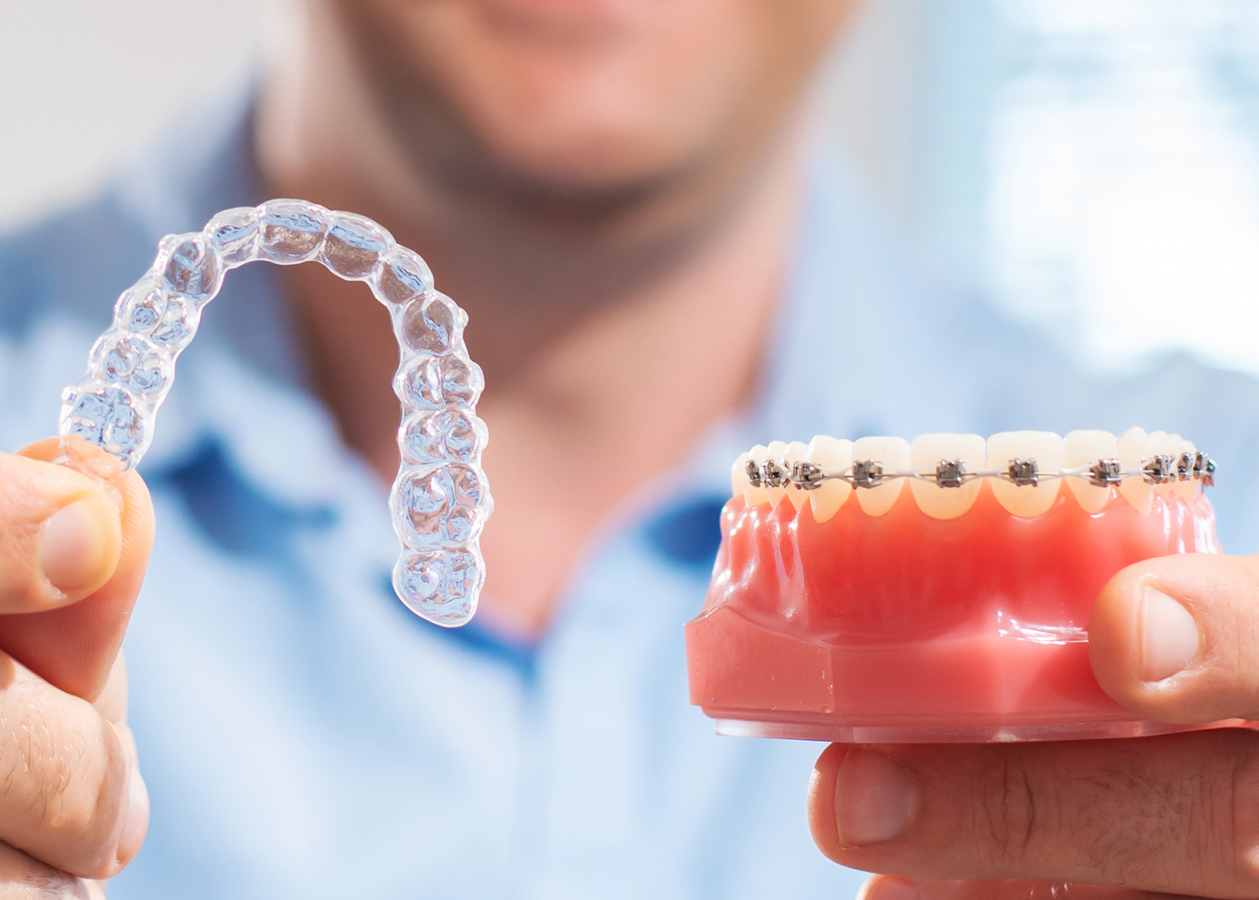 The image shows a person holding up a transparent dental retainer with visible teeth impressions, next to a clear plastic tray with a set of teeth.