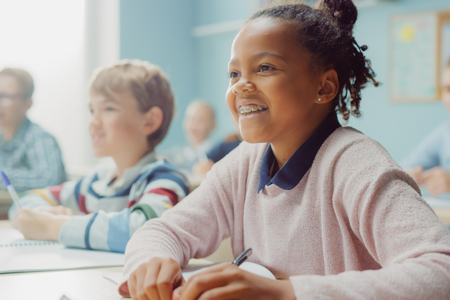 A young girl with curly hair is smiling at the camera while sitting at a desk in a classroom, surrounded by other students who are engaged in their own tasks.