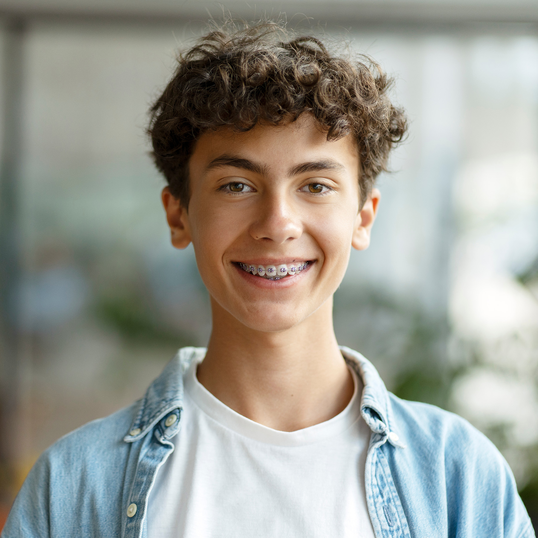The image shows a young male with curly hair smiling at the camera, wearing a light blue denim jacket and standing indoors with a blurred background.