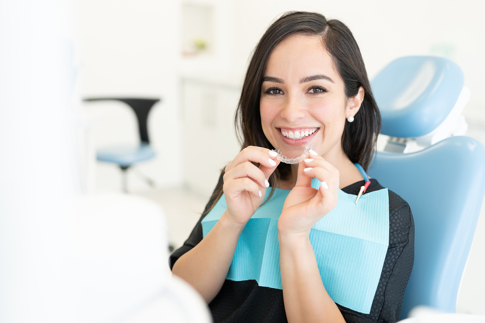 A dental hygienist in a blue chair, holding up a toothbrush with a toothpaste tube, smiling at the camera.