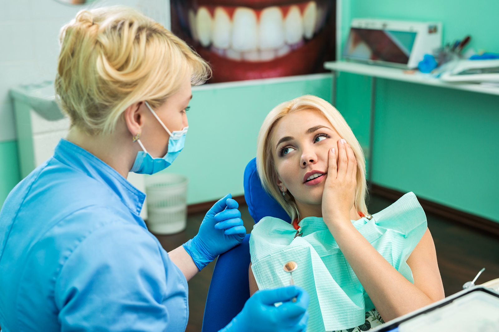 The image shows a dental clinic setting with a woman patient receiving care from a dentist, who is wearing a surgical mask and blue gloves.