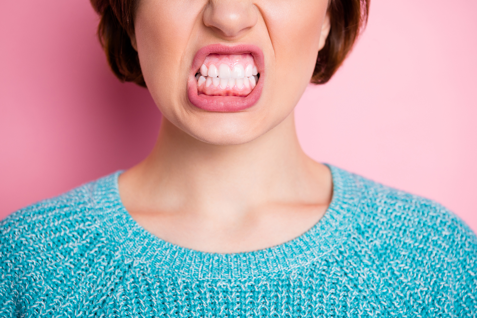 The image features a woman with an exaggeratedly wide-open mouth and teeth, making a face that appears angry or expressing displeasure. She has short hair and is wearing a blue sweater. The background is pink.