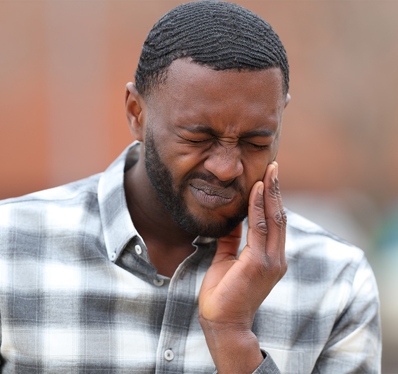 The image shows a man with a hand on his face, possibly rubbing his nose or adjusting his glasses, looking downward with a slight frown, wearing a plaid shirt and a dark-colored cap.