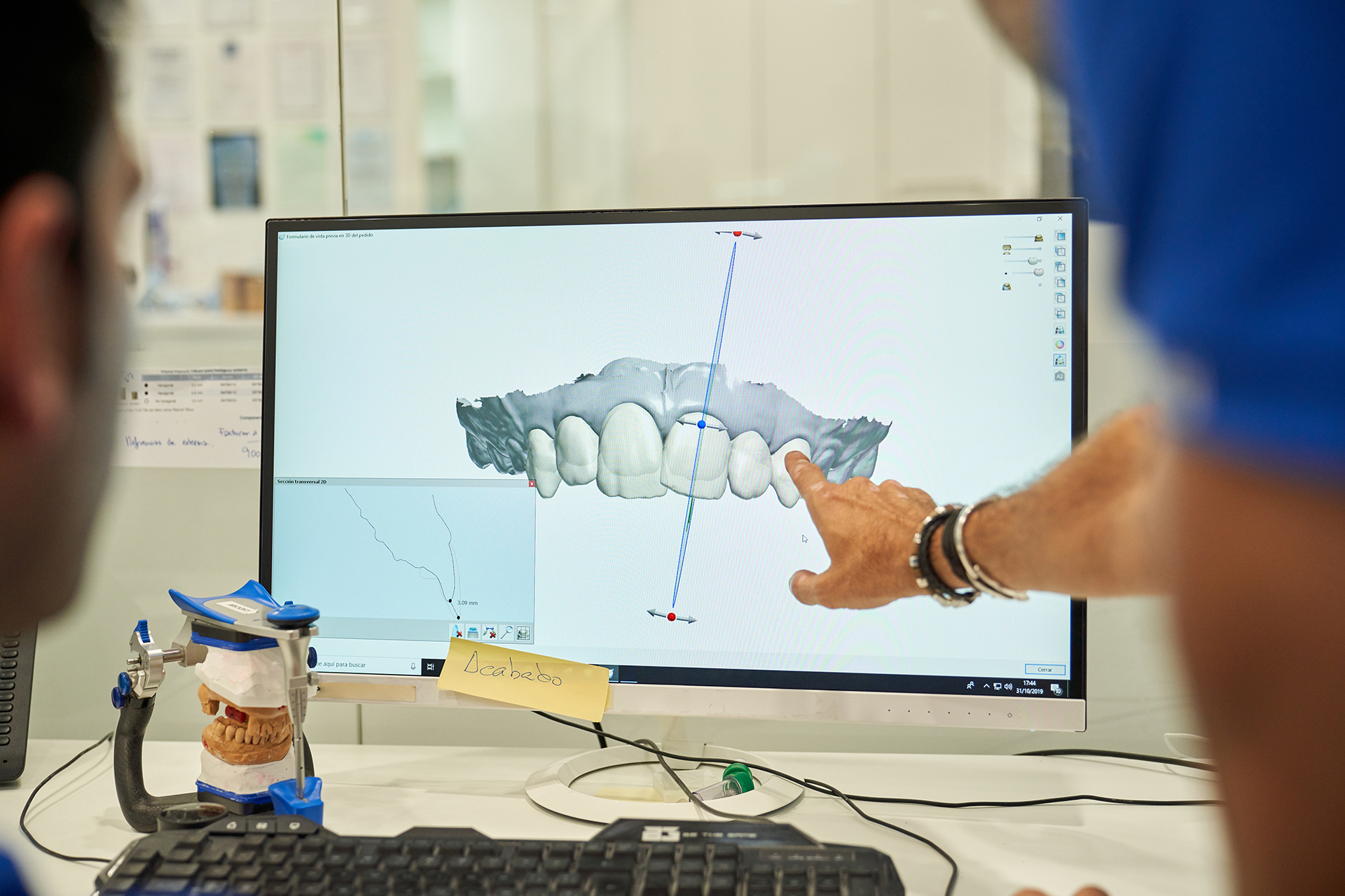 A man and woman are observing a 3D model of teeth on a computer screen, with the man pointing at a specific area, possibly in a dental office setting.