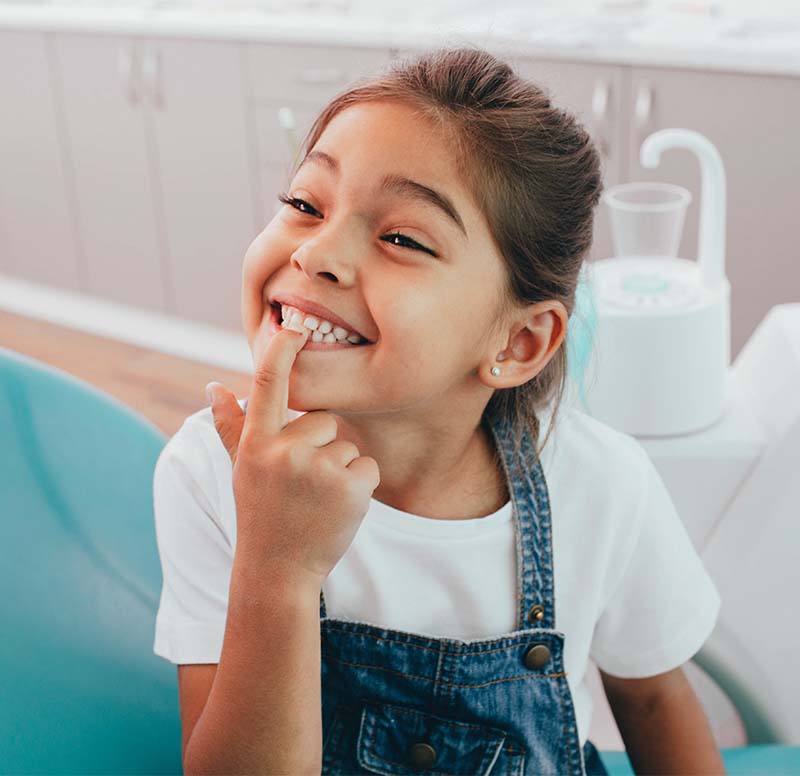 The image shows a young girl sitting in a dental chair, smiling broadly with her finger on her nose, while wearing overalls.