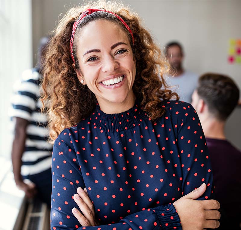 A smiling woman with polka dot shirt stands confidently with hands on hips against office setting backdrop.