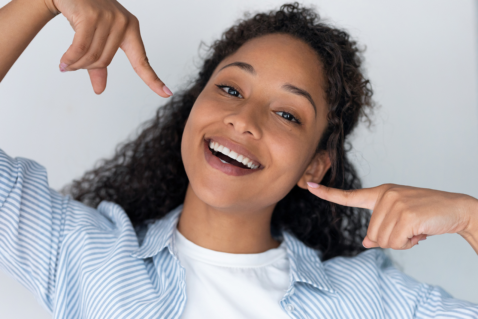 The image shows a person with curly hair smiling broadly while making a peace sign with their hand, standing against a white background.