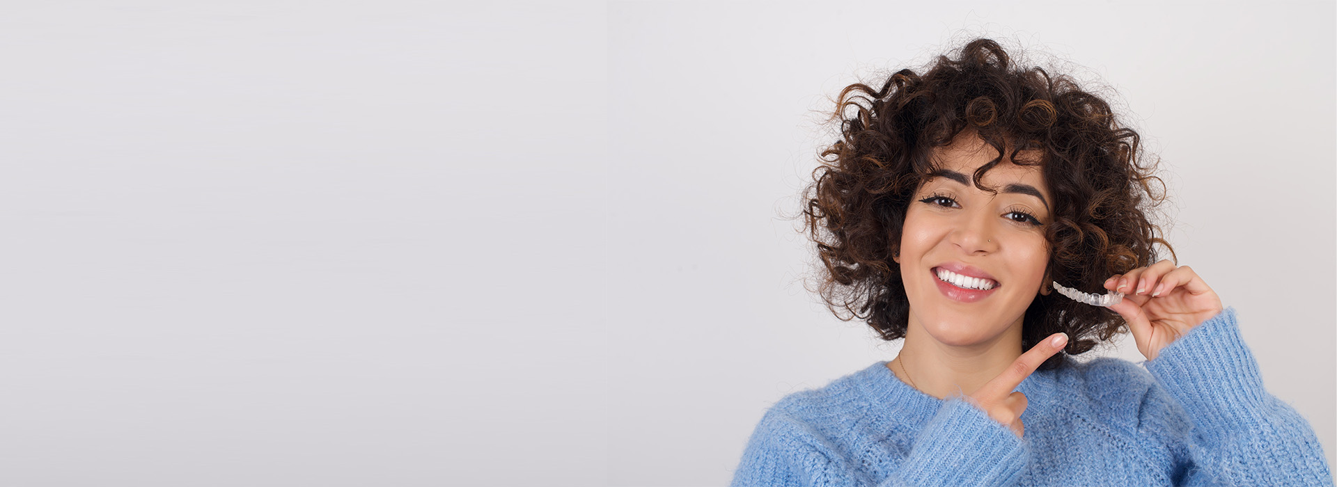 The image shows a person with curly hair smiling at the camera while holding something small in their hand, standing against a plain background.