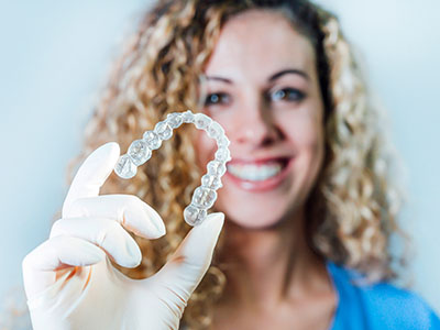 A woman holds up a clear plastic bracelet with a large, round object inside, possibly a dental implant, against her face, smiling broadly.