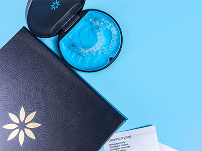 The image shows a dark-colored box with a white flower design on top, placed next to a blue object that appears to be a glass ash tray, all set against a light blue background.