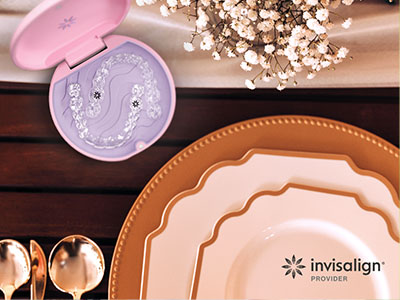 The image shows a table setting with various items such as plates, silverware, a cake stand, a cosmetic case, and a decorative object, all arranged on a wooden surface with a pink lid open in the foreground.