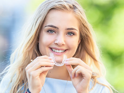 The image shows a young woman with a bright smile holding up a clear plastic mouthguard, displaying her teeth, against a blurred background.