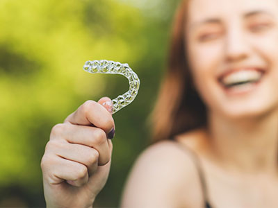 The image shows a smiling person holding up a toothbrush with a smiley face on it against a blurred outdoor background.