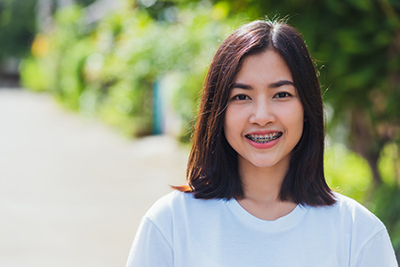 The image shows a person posing for the camera with a smile, wearing a white top, standing outdoors with greenery in the background.