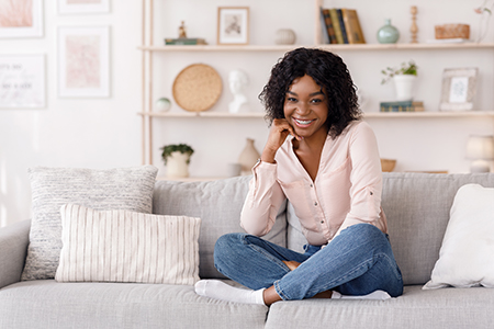 A young woman with dark hair is sitting on a couch, smiling at the camera, wearing a pink top and blue jeans.
