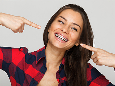 The image shows a woman with a big smile pointing towards her teeth, wearing a plaid shirt and a playful pose.